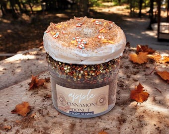 May include: A glass jar filled with brown slime topped with a white frosted donut with cinnamon and sprinkles. The jar has a label that reads "apple cinnamon donut fuzzy slimes".