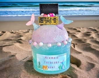May include: A blue and purple slime with a mermaid tail and a treasure chest on top. The slime is in a clear jar with a label that says "Mermaid Treasure Hunt" and "Fuzzy Slimes".