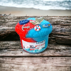 May include: A red and blue slime with a shark, boat, anchor, life preserver, and surfboard on top. The slime is in a clear plastic container with a label that says "Fuzzy Slimes Shark Attack".