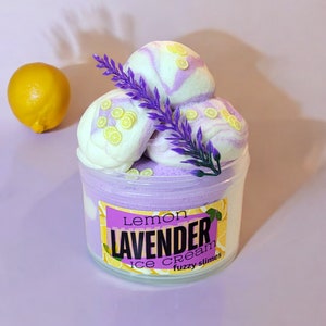 May include: A clear jar filled with purple and white fluffy slime with a label that reads "lemon LAVENDER ice cream fuzzy slimes". The slime is topped with three white and purple swirls with yellow lemon slices on top. A sprig of lavender is placed on top of the slime.