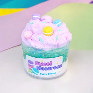 May include: A clear plastic jar filled with a light pink fluffy slime topped with pastel-colored macarons and green sprinkles. The jar has a white label that reads "Sweet Macaroon Fuzzy Slimes".