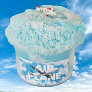 May include: A clear plastic container filled with light blue textured slime topped with a small white airplane figurine. The label on the front features a graphic of an airplane in the sky with the text AIR PLANE and smaller text below.