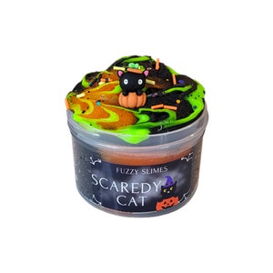 Puede incluir: Una slime negra y verde con un gato negro que lleva un sombrero de bruja y una calabaza en la parte superior. La slime est&aacute; en un recipiente transparente con una etiqueta negra que dice "Fuzzy Slimes Scaredy Cat".
