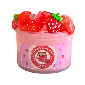 May include: A clear plastic container filled with pink slime, topped with red strawberry-shaped decorations and glitter. The container has a label that reads "Strawberry Jam" and "Fuzzy Slimes."