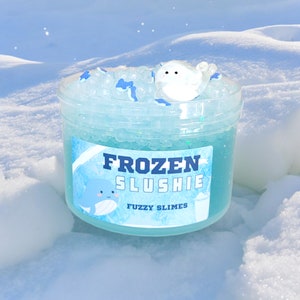 May include: A clear plastic container filled with blue slime, small clear beads, and blue whale-shaped decorations. The label reads "FROZEN SLUSHIE" with a cartoon whale illustration. A white bird-shaped charm sits on top. "FUZZY SLIMES" is printed below.