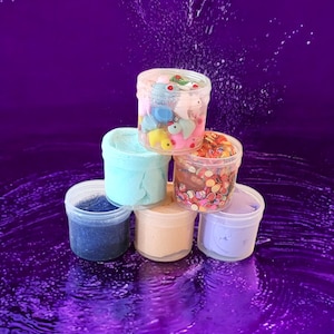 May include: A stack of six clear plastic containers filled with different colored slime. The slime is topped with various colorful decorations, including small toys, sprinkles, and glitter.