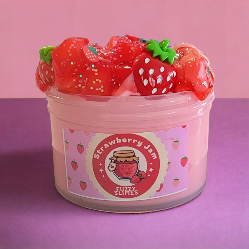 May include: A jar of pink slime topped with red strawberry-shaped decorations and glitter. The jar has a label that reads "Strawberry Jam" with a jar of jam illustration and the words "Fuzzy Slimes". The background is pink and purple.