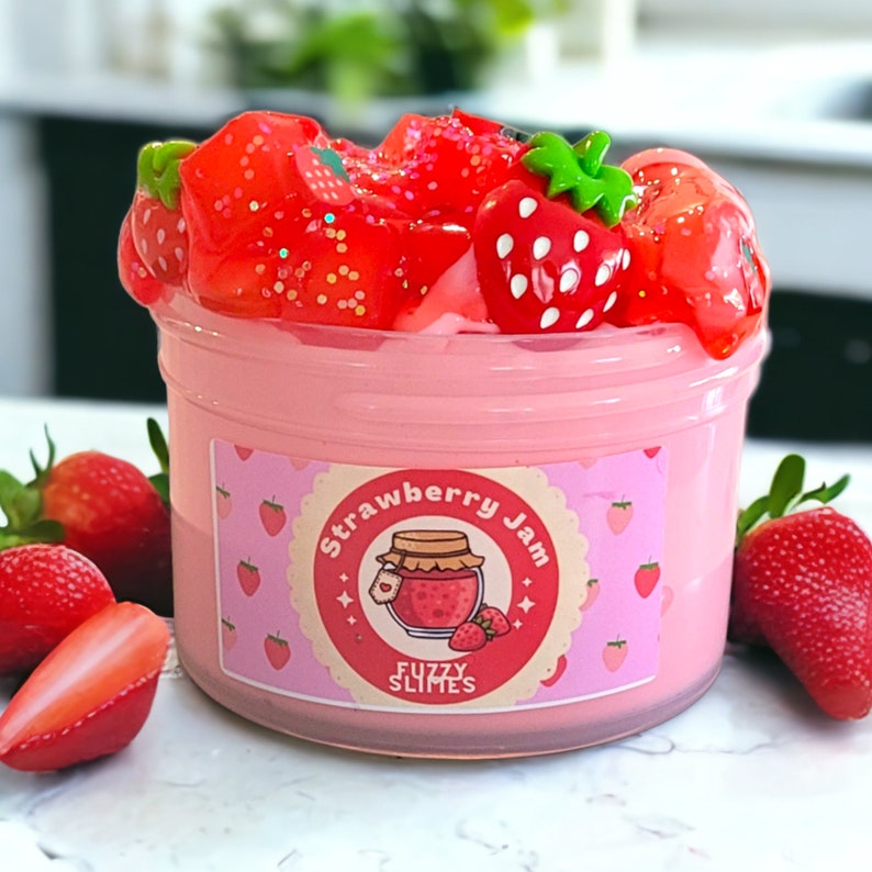 May include: A clear plastic container filled with pink slime, topped with red strawberry-shaped decorations and glitter. The container has a label that reads "Strawberry Jam" with a jar and strawberry illustration. Fresh strawberries are scattered around the container.