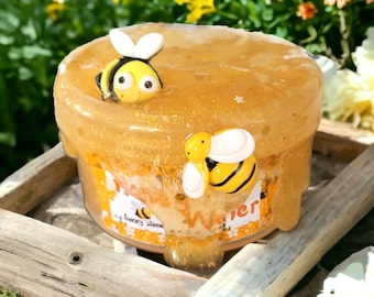 May include: A clear jar filled with honey-colored slime with gold glitter. Two yellow and black bee figurines are partially submerged in the slime. The jar has a label that reads "Honeycomb" and "My Fuzzy Slime".