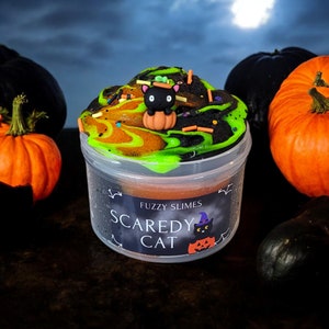 Puede incluir: Un recipiente de pl&aacute;stico transparente lleno de slime verde, naranja y negro con una figurita de gato negro en la parte superior. El recipiente tiene una etiqueta que dice "Fuzzy Slimes Scaredy Cat".