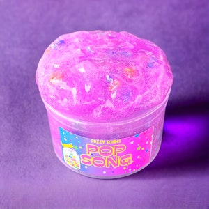 May include: A clear plastic container filled with pink glitter slime with a label that reads "Fuzzy Slimes Pop Song".