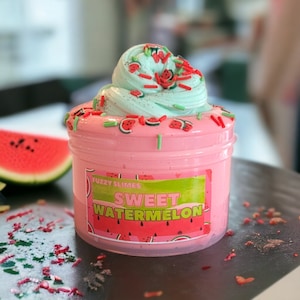 May include: A pink and green slime with watermelon slices and green sprinkles. The slime is in a clear jar with a label that says "Fuzzy Slimes Sweet Watermelon".