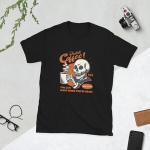 May include: Black t-shirt with a vintage-style graphic of a skeleton drinking coffee. The graphic says "Drink Coffee!" and "You can sleep when you're dead!"