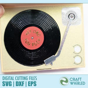 Puede incluir: Una tarjeta dorada con forma de tocadiscos vintage. El disco negro tiene el texto "Hey Dad You Rock" en una etiqueta roja. La tarjeta tiene un brazo plateado y dos botones circulares. La tarjeta est&aacute; sujeta por una mano.