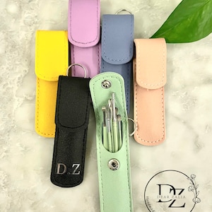 May include: Six different colored leather pouches with a silver keychain. The pouches are lined up in a row. The pouch in the center is open and contains a set of small tools. The pouch is light green and has the initials "D.Z." on it. The other pouches are yellow, black, purple, blue, and pink.