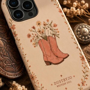 Könnte beinhalten: Eine cremefarbene Handyhülle mit einer zentralen Illustration von zwei rosafarbenen Cowboystiefeln, die mit orangefarbenen Wildblumen gefüllt sind. Ein zarter Blumenrankenrand umrahmt das Design. Der Text DUSTBYJO WESTERN SOUL steht unten rechts.