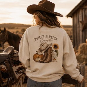Peut inclure: Un sweat-shirt beige à manches longues avec un motif au dos portant le texte PUMPKIN PATCH Cowgirl. Le dessin représente un chapeau de cowboy, des bottes en cuir marron, un tournesol, deux citrouilles et un gobelet marqué PUMPKIN LATTE.