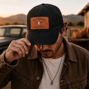 Peut inclure: Casquette de baseball noire avec un patch en cuir portant le texte "DUSTBYJO" et un motif de crâne de taureau. La casquette est portée à l'extérieur, avec un coucher de soleil en arrière-plan. La casquette fait partie de la collection Western Soul.