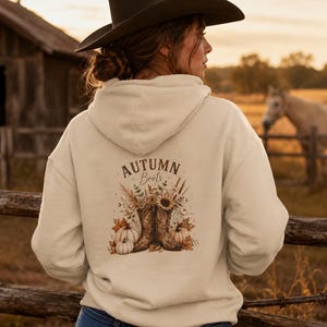Peut inclure: Un sweat-shirt à capuche beige avec un motif au dos portant l'inscription "AUTUMN Boots" au-dessus d'une illustration de deux bottes de cow-boy, tournesols, blé séché et citrouilles blanches, porté avec un jean bleu et un chapeau de cow-boy marron.