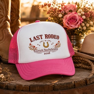 Peut inclure: Une casquette de type trucker rose et blanche avec une impression graphique portant le texte "LAST RODEO", "Emma's Bachelorette" et "2026". Le motif comprend un fer à cheval central entouré d'éléments floraux et de petites étoiles.