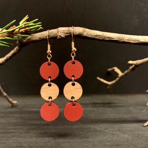 May include: Earrings with a modern design. Each earring features three circular elements: two terracotta-colored discs sandwiching a natural wood disc. The earrings hang from gold-colored hooks, creating a minimalist and stylish accessory.