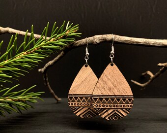 May include: Two wooden teardrop earrings with etched geometric designs. The earrings hang from silver hooks. The background is dark, with a branch and a sprig of evergreen. The earrings are a natural wood colour.