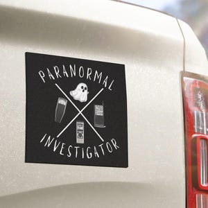 May include: Black square sticker with the words "PARANORMAL INVESTIGATOR" in white, featuring crossed tools and a ghost illustration. The sticker is on the rear of a white vehicle.