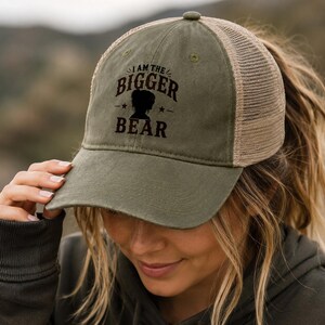 May include: An olive green baseball cap with a tan mesh back and a curved brim. The front panel features dark brown text that reads I AM THE BIGGER BEAR, accompanied by a silhouette of a bear head and two small stars on either side of the graphic.