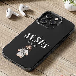 May include: Black phone case with the text "JESUS is King" and a cartoon illustration of Jesus. The case is on a wooden surface with a laptop, earbuds, and a small plant in the background.