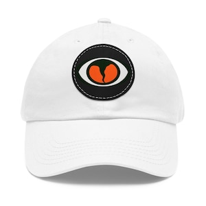 May include: White baseball cap with a black and white eye patch design with an orange iris. The eye patch has a stitched border.