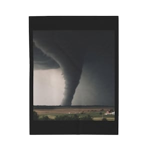 May include: A blanket featuring a dramatic image of a tornado touching down in a rural landscape. The swirling dark gray funnel cloud dominates the scene, contrasting with the green and brown fields below. The blanket is framed in black.
