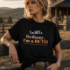 May include: A black short-sleeved t-shirt featuring the white and orange text: I'm NOT a Bethany. I'm a BETH. I am the rock therapists break themselves against. The shirt has a crew neckline and rolled sleeves, worn with a brown leather belt.