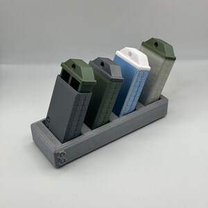 Peut inclure: Un porte-revues imprimé en 3D de couleur grise avec quatre magazines. Les magazines sont de différentes couleurs : noir, vert, bleu clair et gris.