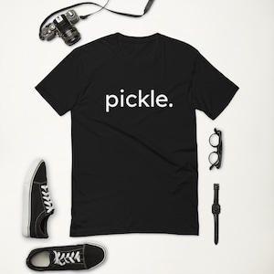 Puede incluir: Camiseta negra con la palabra "pickle." impresa en blanco en la parte delantera. La camiseta está colocada sobre una superficie blanca con una cámara, gafas, un reloj y dos pares de zapatos negros.