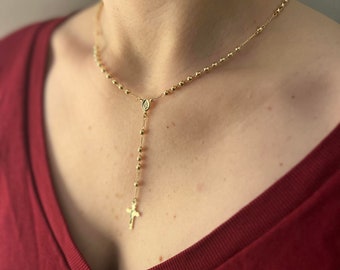 Puede incluir: Un collar de rosario dorado con un pequeño colgante de cruz y un dije con la Virgen María. El collar lo lleva una persona con una camisa roja.