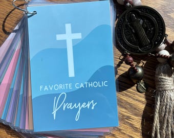 Könnte beinhalten: Ein Stapel Gebetskarten mit blauem Umschlag, weißem Kreuz und der Aufschrift "Favorite Catholic Prayers". Ein Rosenkranz mit Holzperlen und einem Metallanhänger ist ebenfalls zu sehen.