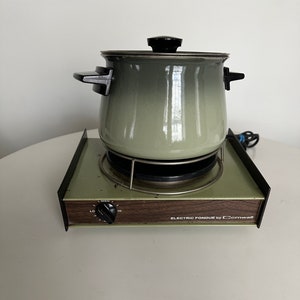 Może przedstawiać: Elektryczny garnek do fondue w stylu vintage, w kolorze zielonym i czarnym, z drewnianą podstawą i pokrętłem do regulacji temperatury. Podstawa ma napis "ELECTRIC FONDUE by Cornwall".