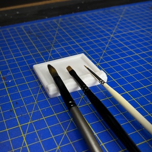 May include: Three paintbrushes of different sizes and colours are resting in a white plastic brush holder. The holder has three slots for brushes and a small rectangular well for water or paint.