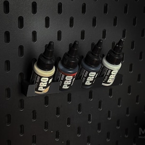 May include: Four bottles of PRO paint are displayed on a black pegboard. The bottles are black with white and red labels. The pegboard has a series of evenly spaced, oval-shaped holes. The bottles are held in a black shelf.