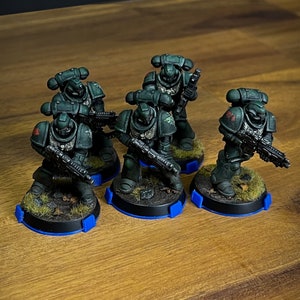 May include: Five green and black painted miniature figures of space marines standing on round bases with blue edges. The figures are holding weapons and are posed in a variety of action poses.