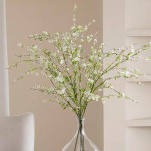 May include: A clear glass vase with a wide base and a narrow neck, filled with white artificial flowers with green stems.