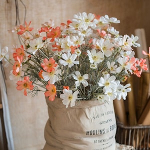 May include: A bouquet of white and peach-colored flowers in a burlap sack with the text "Sac en Location Grains Moulins de Bouv 67 Avenue de Marc" printed on the side.