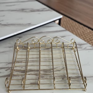 Pode incluir: Um suporte de metal dourado com formato retangular. O suporte apresenta uma série de barras verticais e detalhes decorativos na parte superior e nas laterais. Ele foi projetado para segurar itens e é colocado em uma superfície clara.