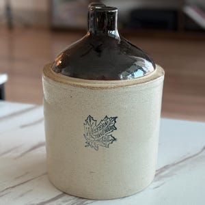 May include: A vintage stoneware jug with a dark brown glazed top and handle. The cylindrical body is a light beige color with a black and white logo that reads "Western Stoneware". This antique jug is a decorative piece.