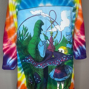 Op de afbeelding: Een tie-dye T-shirt met een kleurrijk psychedelisch ontwerp met Alice in Wonderland en de rups die een waterpijp rookt op een paddenstoel.