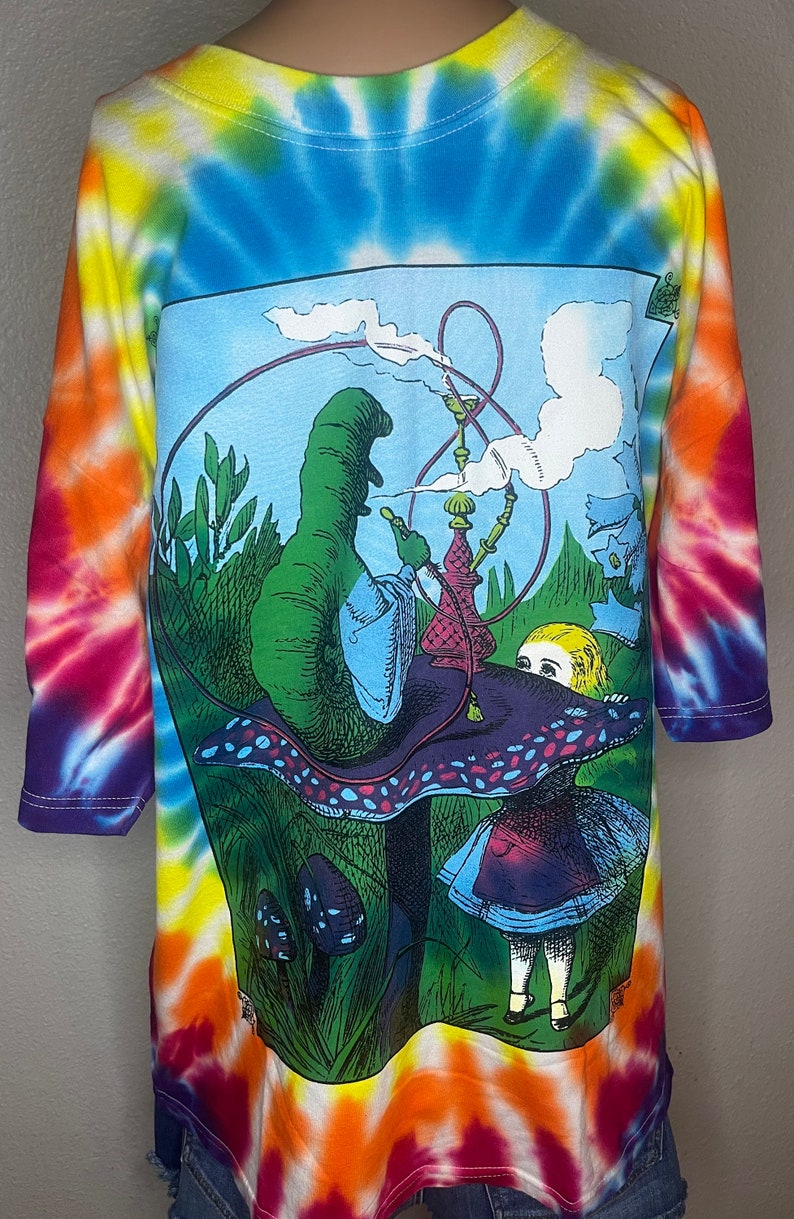 Op de afbeelding: Een tie-dye T-shirt met een kleurrijk psychedelisch ontwerp met Alice in Wonderland en de rups die een waterpijp rookt op een paddenstoel.