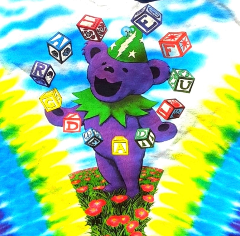 Puede incluir: Una camiseta de color morado con estampado tie-dye con un oso de dibujos animados haciendo malabares con bloques coloridos con letras. El oso lleva un sombrero verde con un rayo y una estrella. El fondo es un patr&oacute;n tie-dye de azul, amarillo y verde.