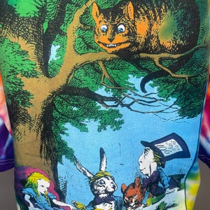 Op de afbeelding: Een tie-dye T-shirt met een kleurrijke illustratie van personages uit Alice in Wonderland. De Cheshire Cat zit op een tak boven Alice, de Hoedenmaker, de Maartse Haas en de Slaapmuis die aan een tafel zitten met een theepot en kopjes.