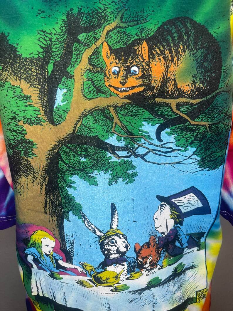 Op de afbeelding: Een tie-dye T-shirt met een kleurrijke illustratie van personages uit Alice in Wonderland. De Cheshire Cat zit op een tak boven Alice, de Hoedenmaker, de Maartse Haas en de Slaapmuis die aan een tafel zitten met een theepot en kopjes.