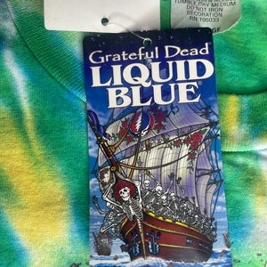 以下が含まれることがあります： スケルトンとバラの船が描かれたタグが付いたタイダイTシャツ。テキストは「Grateful Dead LIQUID BLUE」です。シャツは緑、青、黄色の模様です。